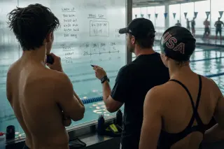 Coach draws swim tactics on glass for focused young swimmers by the pool, early morning, with subtle aquatic reflections and authentic training atmosphere.