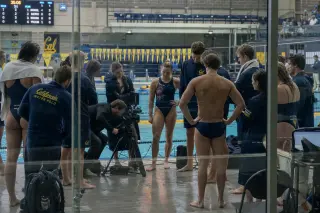 Swimmers and water polo athletes near a poolside media setup, technicians preparing broadcast equipment, documentary-style scene with reflections and authentic ambiance.