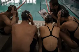 Swimmers in wet gear intently reviewing tactical notes with their coach at the edge of a pool, subdued natural light, candid and focused mood.