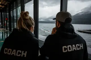Coaches in indoor pool observe open water swimmers training in stormy outdoor lake through large windows, illustrating contrast between indoor and outdoor aquatic environments.