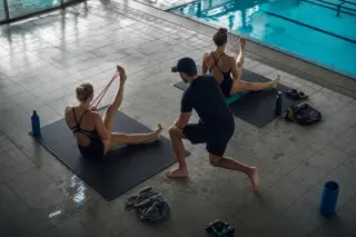 Elite swimmers and coach stretching with resistance bands on poolside mats during early morning preparation, natural light reflecting off the pool.