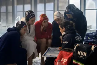 Female water polo players and coach huddled beside the pool, studying a tactical board in soft natural light, showing focus and team camaraderie.