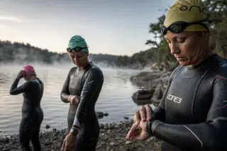 Swimmers in wetsuits adjusting equipment on a misty lakeshore at dawn, focused and preparing quietly by the water