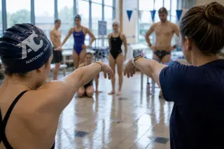 Coach guiding a young swimmer's arm movement in a dryland drill by the pool, with team stretching in background, calm natural light and a focused atmosphere.