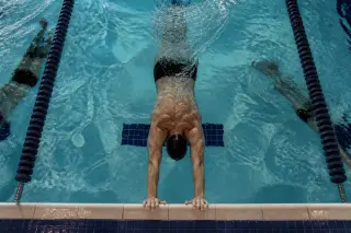 Swimmer's hands gripping the pool edge underwater, captured from above with subtle natural light and a focused, documentary style.