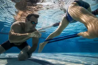 Underwater view of a coach and swimmer analyzing leg kick technique with resistance band in a pool, seen from below with natural light and realistic reflections.