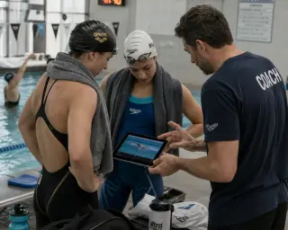 Adult swimmers and coach reviewing video analysis by the poolside, focused expressions, natural pool lighting, candid moment