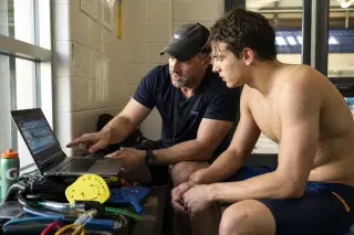 Coach and swimmer in technical room by the pool reviewing swim footage with training equipment nearby, bathed in natural light and pool reflections.