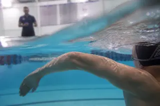 underwater close-up of swimmer's arm in backstroke, coach observing from poolside, technical focus