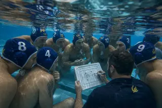 Underwater view of a water polo team and coach in a tactical huddle, all partially submerged, focused on a strategy board, with sunlight filtering through pool water.