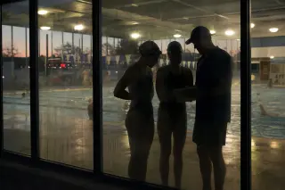 Coach and two swimmers reflect through misted pool glass, consulting notes in focused, natural poolside talk at dusk.
