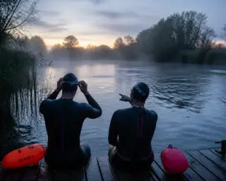 Two open water swimmers adjusting gear on a riverside pontoon at dawn, surrounded by mist and natural elements