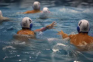 Two water polo players transitioning in the pool, low angle from poolside with focus on hand movement and water droplets, teammates blurred in the background under natural pool lighting.