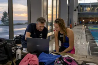 Coach and swimmer in damp gear reviewing swim footage on a laptop beside a pool at dusk with subtle, natural lighting and focused expressions.