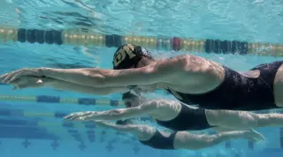 Underwater view of a synchronized group of swimmers in tight formation, focused and streamlined during a training drill, with sunlight filtering through real pool water.
