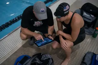 Coach and swimmer analyzing race strategies with a digital tablet by the poolside, dimly lit with authentic Olympic pool ambiance.