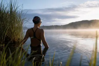 A solitary open water swimmer stands still in a misty lakeside at daybreak, partly submerged, attentively soaking in the atmosphere, with soft natural light and tranquil surroundings.