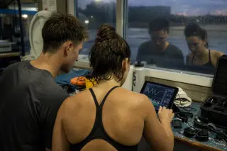 Two swimmers in training attire studying connected swim goggles and wrist sensors at a workbench in a pool equipment room, natural evening light creating an authentic, concentrated mood.