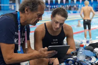 Coach and young swimmer reviewing race data on a tablet poolside, with blurred competition in the background under natural pool lighting.